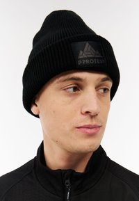 Zwarte gebreide beanie met een geribbeld patroon en een zwart logo-opdruk met een geometrisch ontwerp en tekst. Gedragen met een zwarte ritsjack.