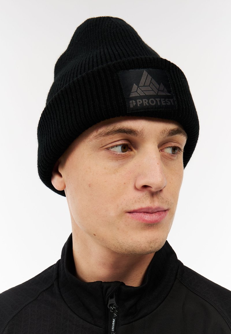 Zwarte gebreide beanie met een geribbeld patroon en een zwart logo-opdruk met een geometrisch ontwerp en tekst. Gedragen met een zwarte ritsjack.