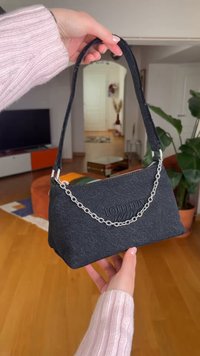 Sac d’épaule noir Tommy Jeans avec détail chaîne argentée, tenu par des mains portant des manches tricotées rose clair dans un salon.