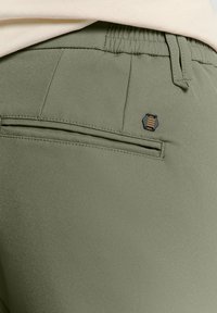 Olivgrüne, schmal zulaufende Hose mit einem elastischen Bund und einer Gesäßtasche, die mit einem hexagonalen schwarz-goldenen Logo-Akzent versehen ist. Glatte Textur.