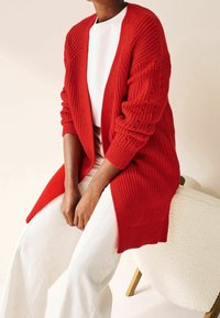 Cardigan rosso lavorato a maglia con texture a coste, apertura frontale e maniche lunghe, abbinato a pantaloni bianchi a gamba larga e posizionato su uno sfondo neutro.