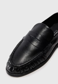 Mocasín de cuero negro con un acabado liso, que presenta una puntera cosida, una correa en la parte superior y detalles recogidos a lo largo de la suela.
