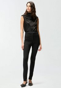 Crni satenski top bez rukava s naboranim izrezom, uparен s crnim hlačama visokog struka koje imaju bočne proreze i glatku teksturu.