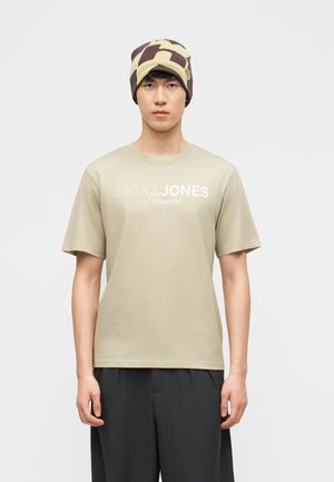 Hombre con camisa beige Jack & Jones, pantalones oscuros y un gorro con patrón marrón y beige, de pie frente a un fondo claro y liso.