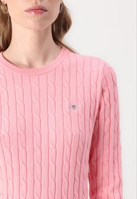 Rosa strikket genser med kabelmønster, rund halsutringning og en liten logo detalj på brystet. Stoffet har en myk tekstur.