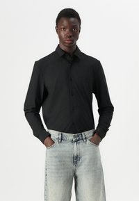 Camisa negra de manga larga con collar tipo botón, con un bolsillo en el pecho, combinada con unos vaqueros de mezclilla sueltos de color azul claro.