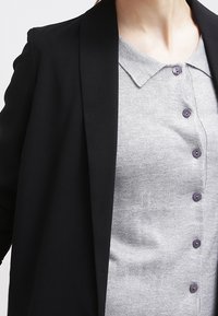 Blazer nero sopra un top a maglia grigio chiaro con colletto e sei bottoni viola, che mette in mostra texture lisce e una vestibilità su misura.
