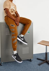 Person som bär en beige Jordan-t-shirt, bruna Jordan-mjukisbyxor och svarta Nike-skor, sitter på ett grått metallskåp i ett rum med blå matta.