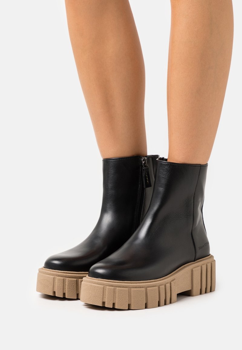 Bottes noires en cuir jusqu'à la cheville avec semelles compensées beiges épaisses portées sur des jambes nues, détail de fermeture éclair sur le côté intérieur de chaque botte.
