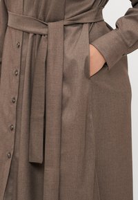 Barna gombos ruha texturált anyagból, derékhoz kötött övvel és oldalsó zsebekkel. Sötét gombok találhatók az elején.