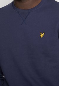 Sudadera azul marino con cuello redondo, costura texturizada y un logo de pájaro bordado en amarillo en el lado izquierdo del pecho. Material de algodón suave.