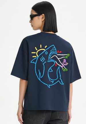 Woman with straight black hair wearing sunglasses and a navy t-shirt showing a colorful shark graphic biting legs with green heels.