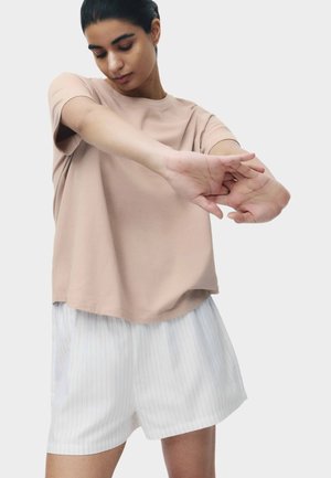 Kobieta w beżowej koszulce z krótkim rękawem i białych szortach w paski rozciągająca ramiona na klatkę piersiową na jednolitym tle.