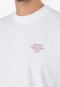 Camiseta blanca de algodón con cuello redondo y mangas cortas. Presenta un bordado rosa con la marca en el pecho: "PEGADOR Certified Authentic Streetwear AWG".