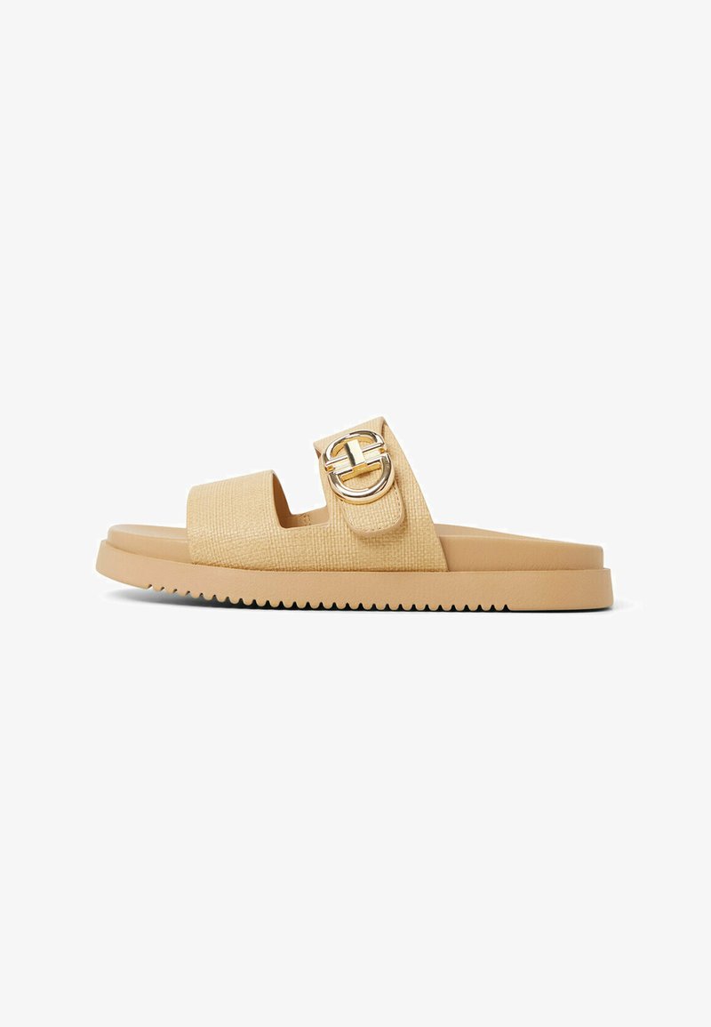 Lys tan slide sandal med en struktureret overdel, en bred rem og en guldfarvet cirkulær spændeaccent. Har en flad gummisål med riller.