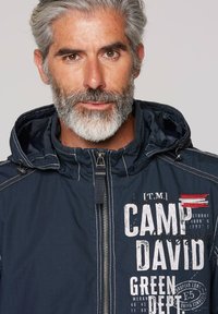 Navy blue zip-up jacket with a hood, featuring white lettering and red stripes as accents. Made of a smooth, lightweight fabric.