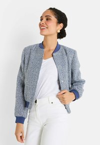 Femme souriante, portant une veste bomber texturée bleue sur un t-shirt blanc et un pantalon blanc taille haute, devant un fond uni.