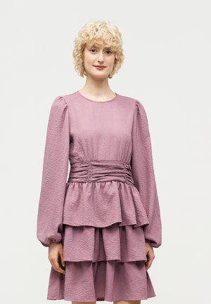 Jeune femme aux cheveux blonds courts et bouclés, portant une robe longue mauve à manches longues et à couches texturées, debout devant un fond uni clair.