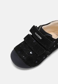 Chaussure noire pour tout-petit avec embout en caoutchouc, deux bandes Velcro, motifs subtils en forme de cœur, et étiquette de marque "GEOX" sur la sangle supérieure.