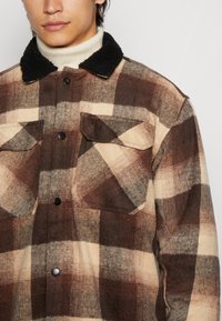 Giacca a camicia a quadri marrone e beige con un colletto in shearling nero. Presenta due tasche sul petto e bottoni lungo la parte anteriore. Tessuto texturizzato.