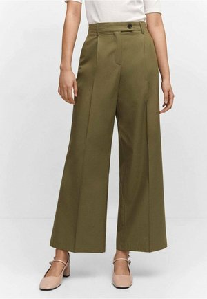 Kobieta ubrana w oliwkowozielone szerokie spodnie z wysokim stanem i plisami z przodu, w połączeniu z beżowymi butami na obcasie typu Mary Jane oraz białą bluzką.