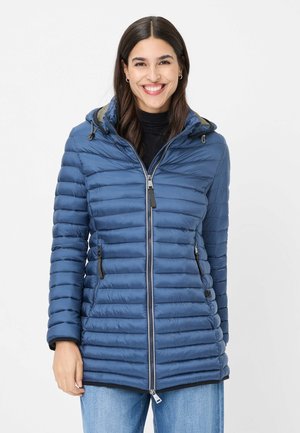 Lächelnde Frau trägt eine blaue gesteppte Kapuzenjacke mit Frontreißverschluss und Reißverschlusstaschen, kombiniert mit hellblauen Jeans.