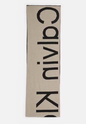 Écharpe beige avec de grandes lettres noires formant verticalement le nom « Calvin Klein » sur toute sa longueur.