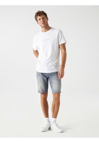 Camiseta blanca de algodón con texto en relieve, combinada con pantalones cortos de mezclilla claros y zapatillas blancas. Atuendo casual sobre un fondo neutro.