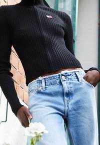 Henkilö, jolla on päällä musta ribbineule ja pieni logo, sekä vaaleansiniset korkeavyötäröiset farkut, seisoo tiiliseinää vasten ja pitää valkoisia kukkia käsissään.