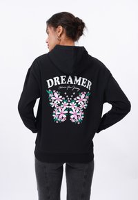 Crna dukserica s cvjetnim uzorkom s ružičastim cvjetovima i zelenim detaljima. Na leđima se nalazi natpis "DREAMER" s porukom "Prihvati svoje putovanje."