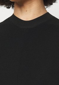 Czarny prążkowany dzianinowy top z wysokim kołnierzem, teksturowaną tkaniną i subtelnymi detalami szwów na przodzie. Zbliżenie na obszar kołnierza.