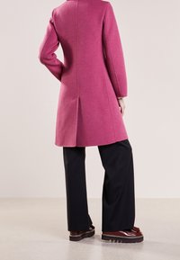 Aansluitend roze wollen jas met lange mouwen en een centrale achterkantventilatie, gecombineerd met zwarte wijde broek en glanzende bruine platform schoenen.