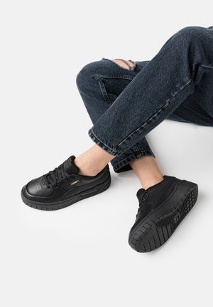 Jambes en jean noir déchiré portant des baskets noires Puma, un pied à plat et l'autre levé montrant le motif de la semelle.