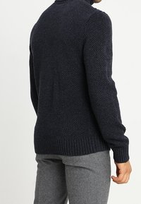 Uomo con un maglione scuro navy a trama e pantaloni grigi, visto di spalle con la mano rilassata al fianco su uno sfondo bianco.
