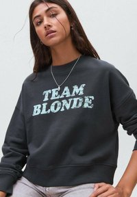 Giovane donna con lunghi capelli castani che indossa un felpa scura con la scritta "TEAM BLONDE" e pantaloni chiari su uno sfondo semplice.