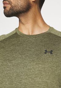 Lähikuva miehen niskasta ja olkapäästä, joka on pukeutunut oliivinvihreään Under Armour -paitaan, jossa on musta logo rinnassa.