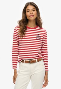 Rood-wit gestreept overhemd met lange mouwen en ronde hals, met een logo patch op de borst. Gepaard met lichtgekleurde broek en een gevlochten riem.