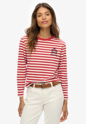 Camicia a maniche lunghe a righe rosse e bianche con scollatura rotonda, caratterizzata da una toppa con logo sul petto. Abbinata a pantaloni chiari e una cintura intrecciata.