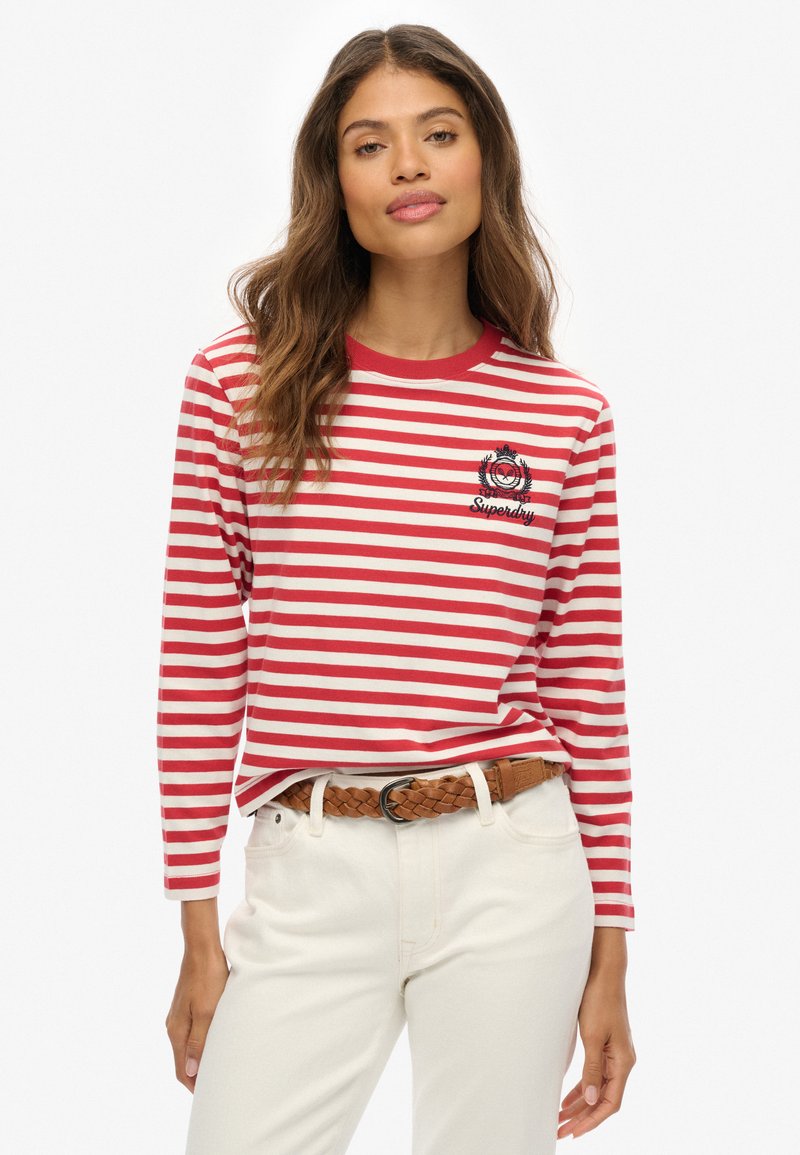 Rood-wit gestreept overhemd met lange mouwen en ronde hals, met een logo patch op de borst. Gepaard met lichtgekleurde broek en een gevlochten riem.