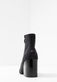 Botín negro hasta el tobillo con una parte superior de ante texturizado, diseño geométrico, puntera redonda y un tacón grueso y brillante. Cuenta con una cremallera lateral y un acento de logo.