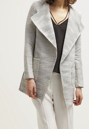 Vrouw die een lichtgrijze, gestructureerde jas met open voorkant draagt over een zwart topje met bandjes en witte wijde broekspijpen, staand tegen een effen achtergrond.