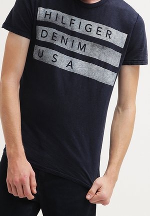 Mand iført en sort T-shirt med teksten "HILFIGER DENIM USA" i grå blokke over brystet, som holder T-shirtens kant med den ene hånd.
