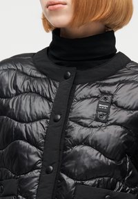 Άτομο με κοντά κόκκινα μαλλιά που φοράει μαύρο μπουφάν με ραμμένα τετράγωνα και πατιλέτα με κουμπιά, καθώς και μαύρο πουλόβερ με ζι βάγκο.