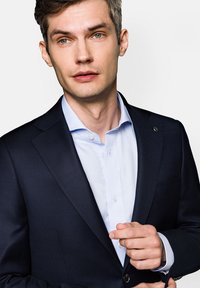 Granatowa marynarka o zwartej konstrukcji, teksturowanej tkaninie i jednym guziku. Jasnoniebieska koszula pod spodem z subtelnym wzorem i standardowym kołnierzykiem.