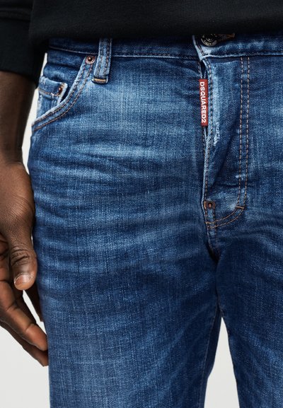 Κοντινό πλάνο σκούρου μπλε τζιν με κόκκινη ετικέτα DSQUARED2 στην εμπρόσθια τσέπη και μαύρο πουκάμισο μερικώς ορατό από πάνω.