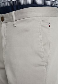 Béžové bavlněné kalhoty s přímým střihem, zapínáním na knoflík, bočními kapsami a malým logem v červené, bílé a modré barvě.