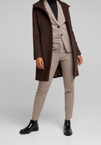 Abrigo marrón sobre un traje de cuadros que presenta una chaqueta ajustada con dos botones y pantalones estrechos. Botines negros completan el conjunto.