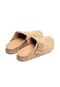Tan kleurige suède klompen met een brede opening, decoratieve metalen studs en een gespaccent. Korken voetbed met een textuur rubberen zool.