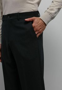 Pantalon gris foncé sur mesure avec une texture lisse, doté d'une fermeture à boutons, de poches latérales et d'un design plat à l'avant.