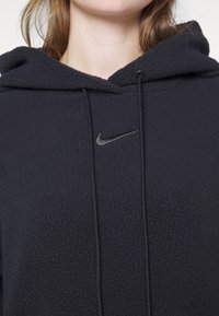 Černá flísová mikina s texturovaným povrchem, přední kapsou a kapucí se šňůrkou. Na hrudi se nachází malé stříbrné logo Nike.